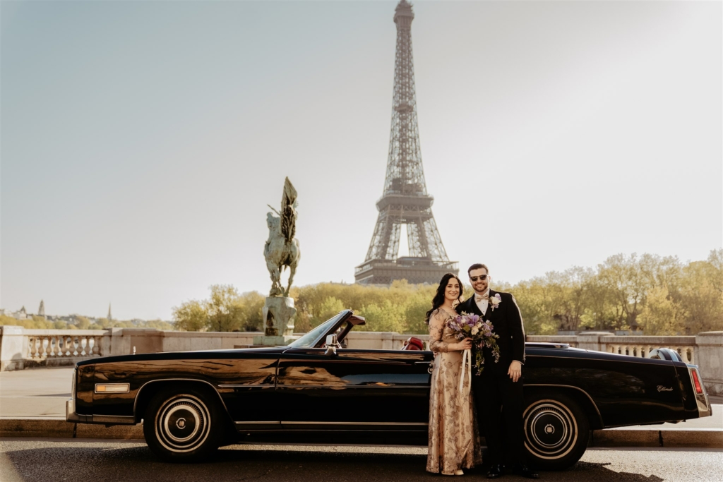 a couple is posing in front of their vintage car at the bir hakeim bridge, with Eiffel Tower views behind, after having their ceremony by a Paris officiant who appears to be english-speaking